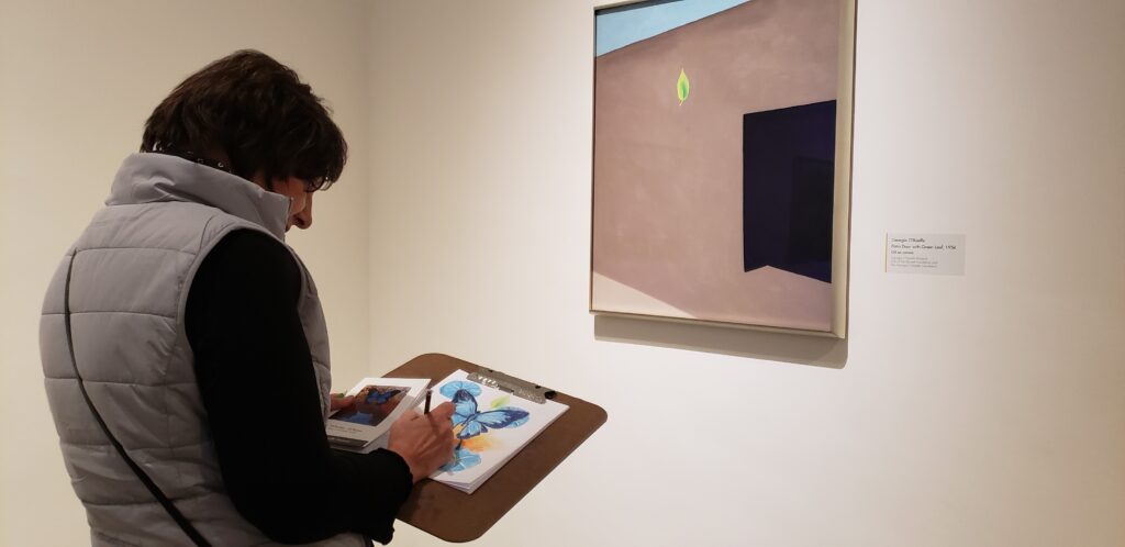 View of a person from the torso up holding a sketch book. They are looking at a painting of Georgia O'Keeffe's adobe patio.