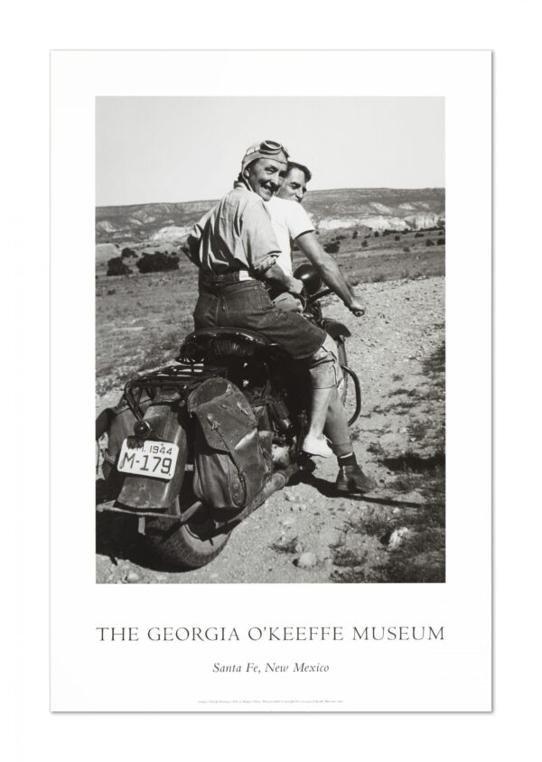 White poster with a black and white photograph in the center and the words 'The Georgia O'Keeffe Museum, Santa Fe, New Mexico' in the bottom. The photograph shows Georgia O'Keeffe sitting behind a companion on a motorcycle. O'Keeffe wear a had cap, goggles, jeans, light shoes and a shirt rolled to her elbows. The motorcycle is on a dirt road and a desert landscape and sky stretches out behind the riders. The motorcycle is turned away and O'Keeffe look back towards the camera. O'Keeffe is smiling.