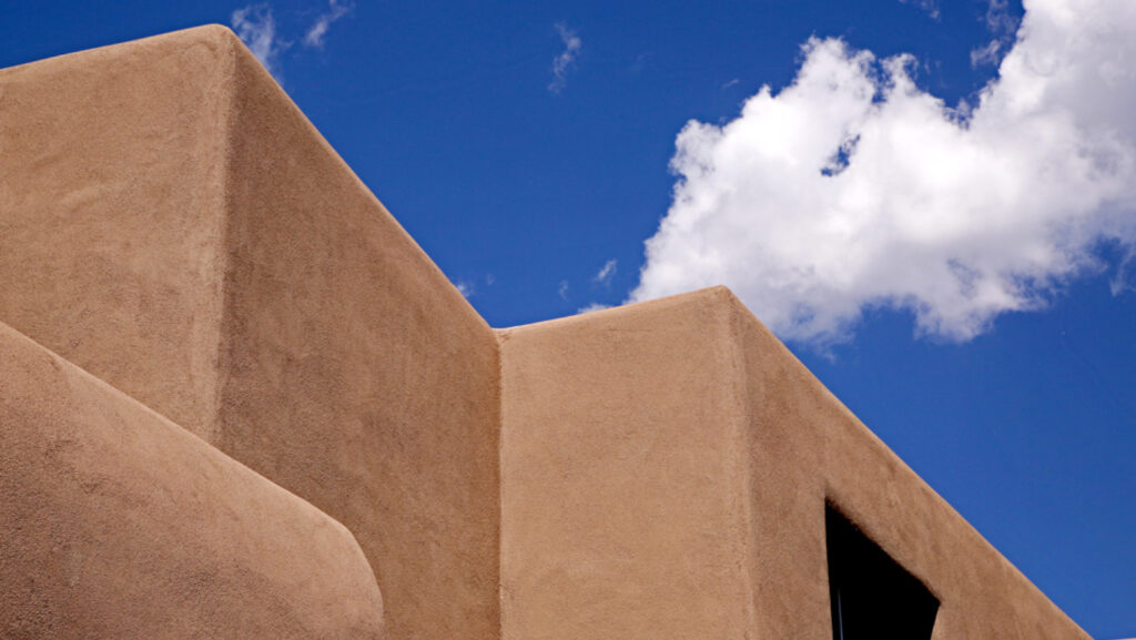 A corner of Georgia O'Keeffe Museum. The image is the top of the adobe colored walls and roof against a brilliant blue sky.