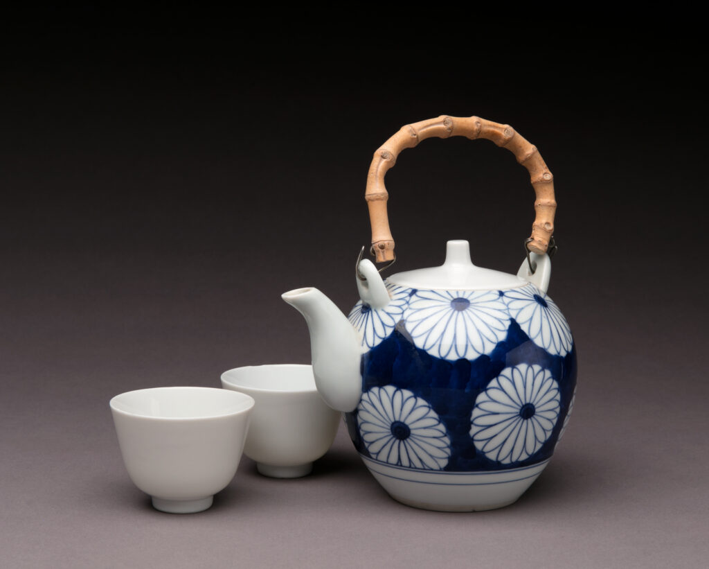 A photograph of a blue daisy floral pattern Japanese teapot with a bamboo handle. On the left are two simple ceramic teacups.