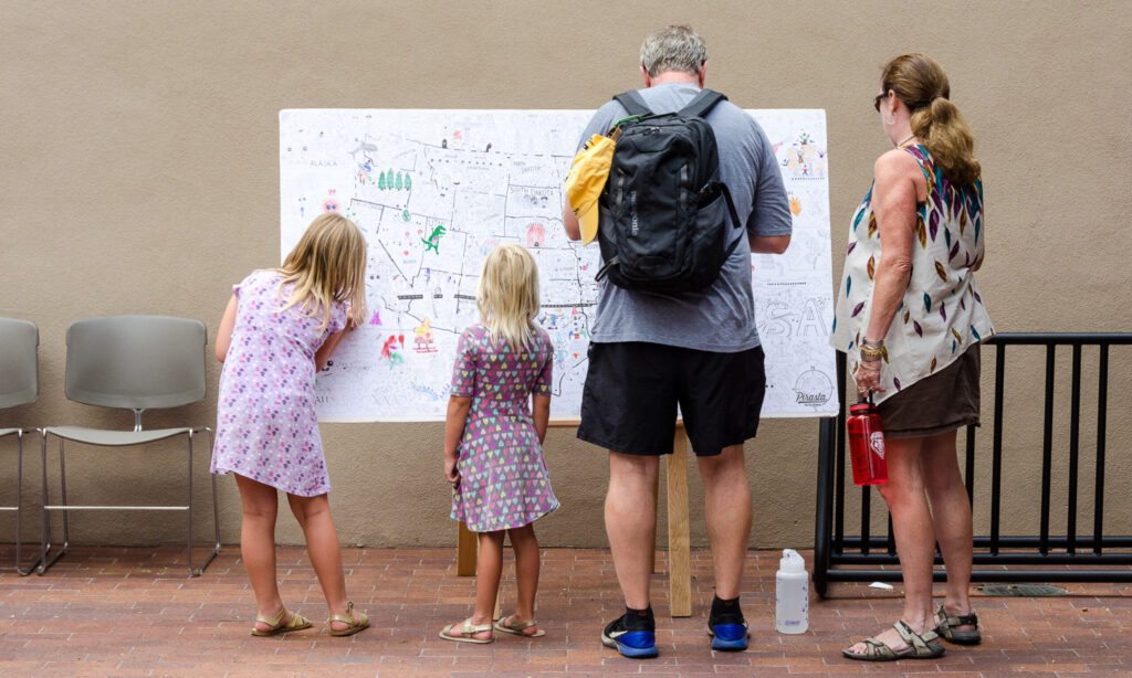 A photograph of two adults and two children taken from behind. They are standing in front of a large whiteboard with a map that has been drawn on by others and the children are contributing to the drawings