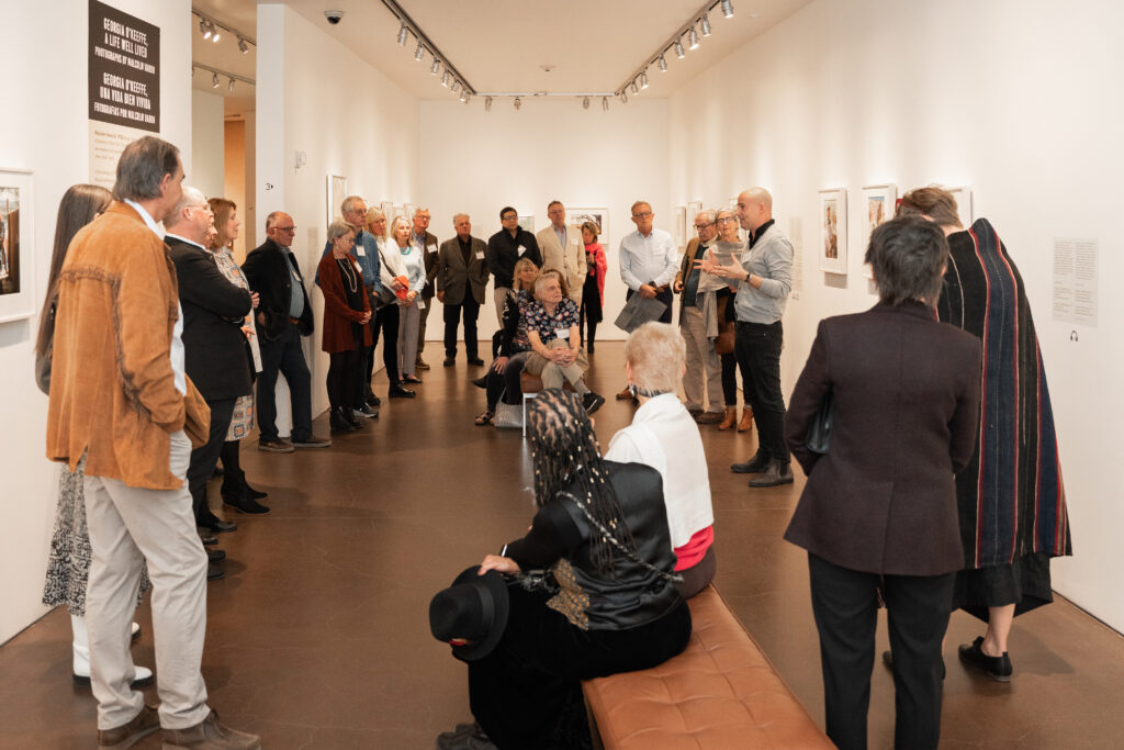 Group of people standing and sitting in a circle listening to a person speak. They are in a museum exhibition space and the person speaking is gesturing with their hands.