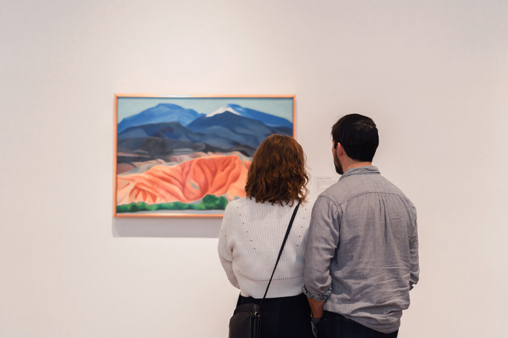 : A photograph of two people taken from behind, they are standing close together looking at O’Keeffe’s painting ‘Black Mesa Landscape, New Mexico / Out Back of Marie's II’ in the Museum galleries.