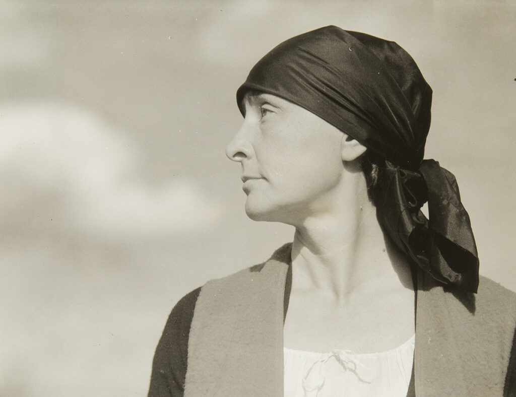 Half-length portrait of Georgia O'Keeffe with head in profile facing viewer left wearing a dark scarf/ turban tied behind her head; torso is frontal, neutral facial expression with eyes looking upward; she wears a white scoop-neck drawstring blouse under a dark sweater and vest; framed against a sky with clouds