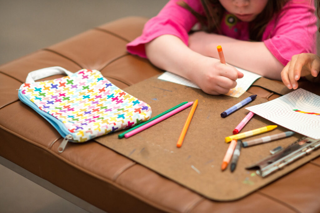 Close-up photograph of a child drawing on a piece of paper with colored pencils on top of a leather bench. The child’s face is out of frame and focus is on their hands, a patterned pencil and the colored pencils scattered over the bench. A second child’s hand pokes in from the right side of the frame.