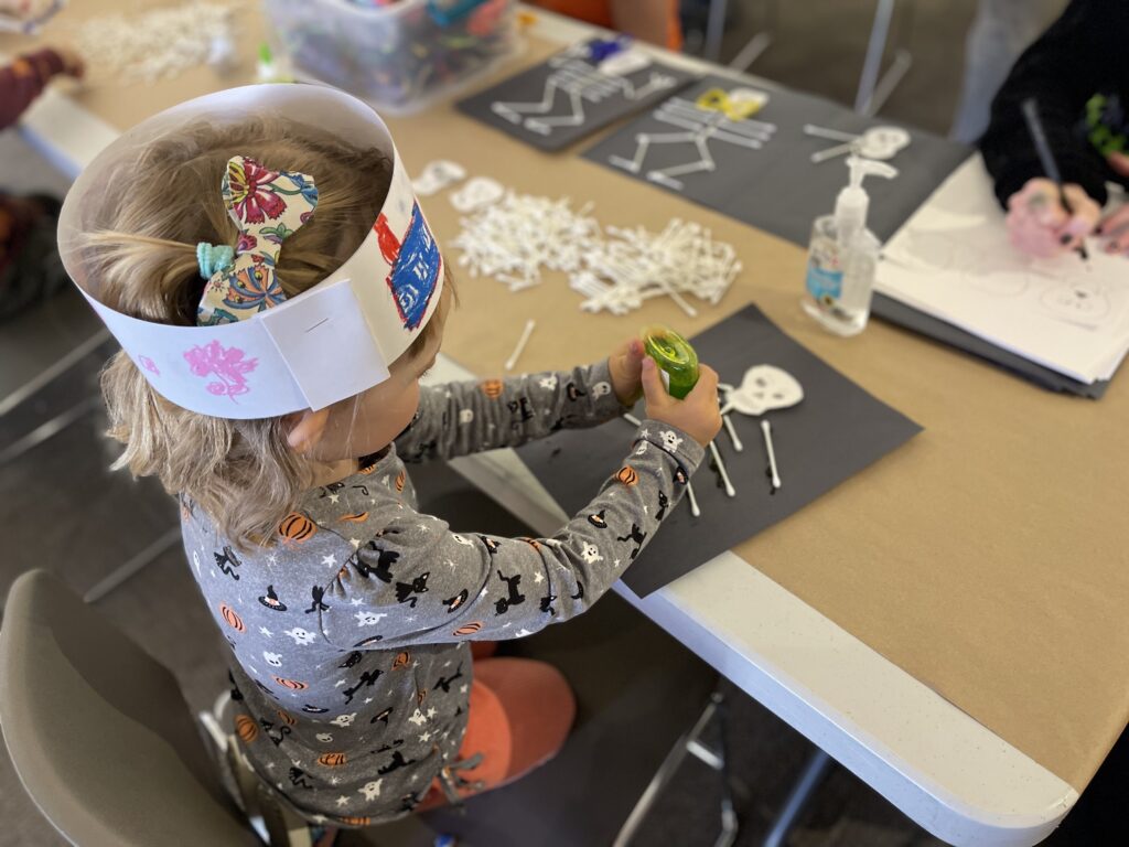 Photo of a child gluing an art piece. The child has a paper crown and the photo is focused on their hands doing the work.