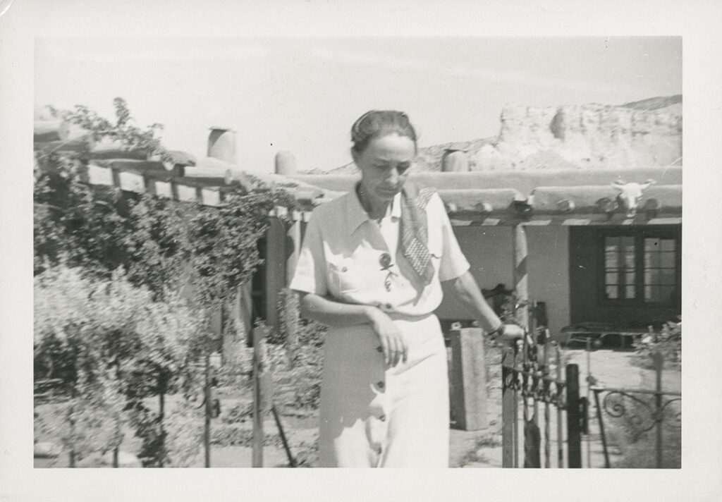 Black and white photograph of O'Keeffe wearing a light buttoned shirt and skirt, her 'OK' pin, and her hair tied up walking through a gate in the Ghost Ranch Patio. Behind her is visible the patio and a skill mounted between Vega beams with the mountains in the background.