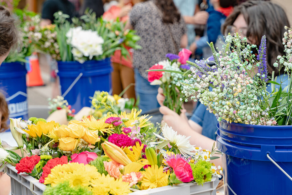 Image of a bunch of bouquets of flowers in containers and buckets on a table. In the background is barely visible a person arranging the flowers