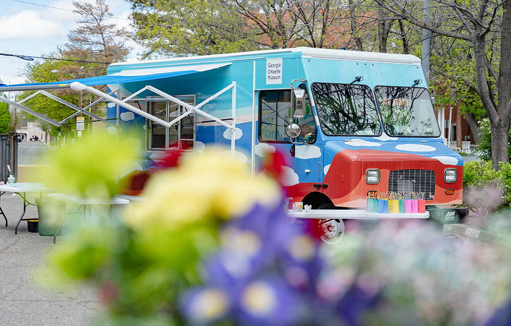 Photograph of the 'Art to G.O.' truck with cloud motifs on the side in a parking lot under trees. the bottom of the frame and front of the truck are partially covered with our of focus/ blurry flowers.