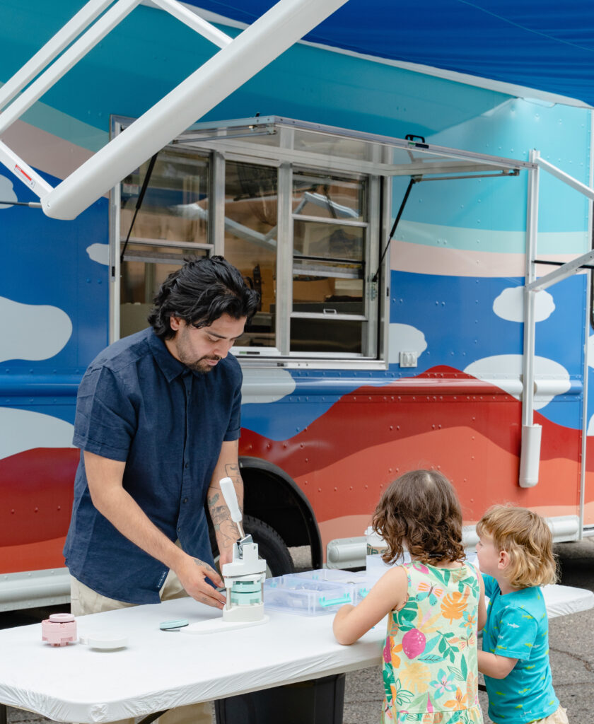 Photograph of a person standing over a table demonstrating making buttons across from two smaller children. Behind them is the Art to G.O. truck with clouds motifs on in.