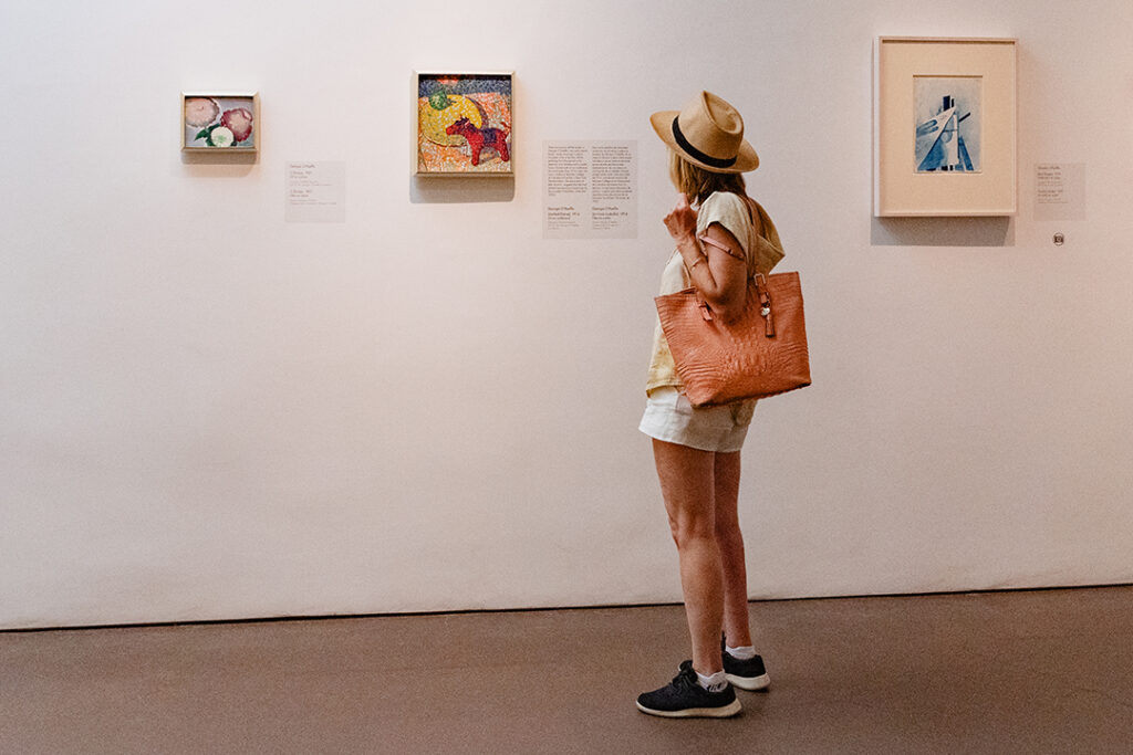 Photograph of a person in a hat and holding a bag looking at three small paintings on a white gallery wall.