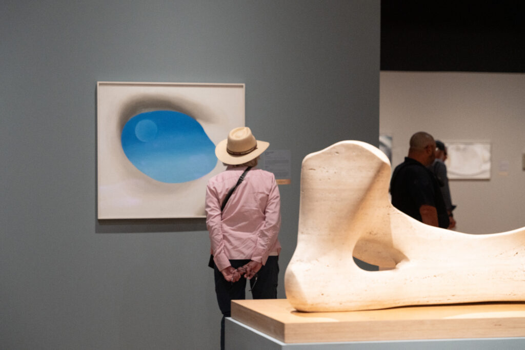 Photograph of a person in a hat from behind standing in front of O'Keeffe's painting of a blue sky and moon through a white pelvis bone. Behind the person is a white sculpture made by Henry Moore lying on a pedestal.