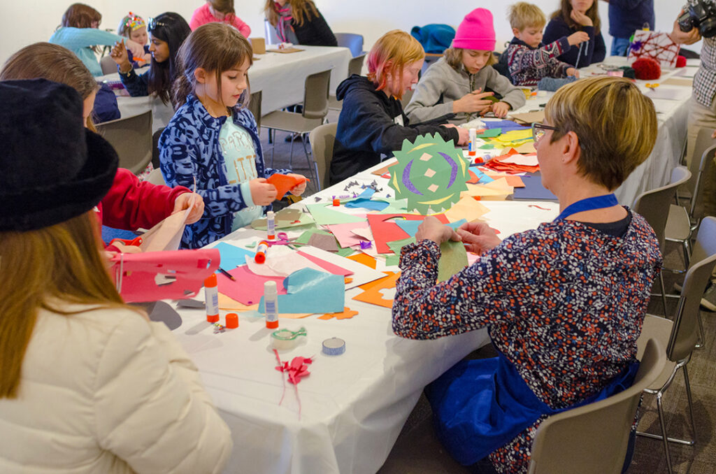 Photograph of a long table with several children and a few adults making art pieces with colorful paper.