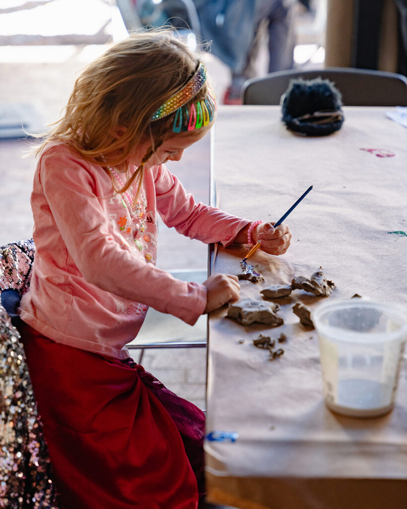 Photograph of a child sitting at a table making art with pieces of clay and a paintbrush.