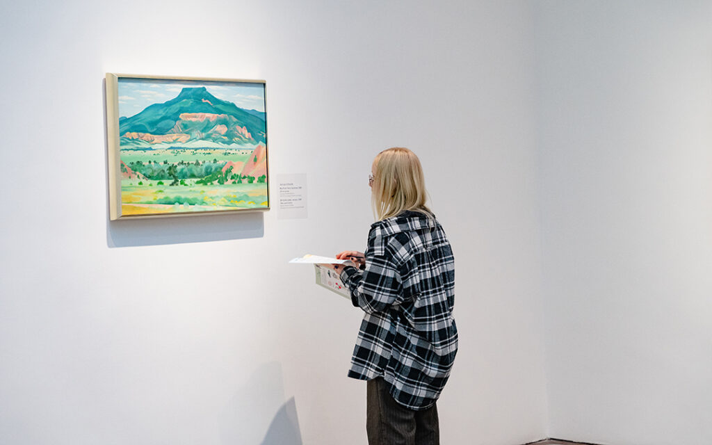 A young person with long hair and glasses holding a paper and pen and looking at a painting on a white gallery wall. The oil painting by O’Keeffe is of a flat-topped mountain in fall, predominantly in dark greens and oranges.