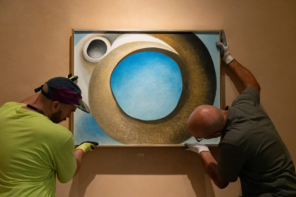 Photograph of two people wearing gloves holding up an art piece onto a beige wall. The person on the left is wearing a hat. The piece by Georgia O'Keeffe is a pastel of a large circular brown goat’s horn. The negative space in the center of the horn and around it are blue. To the left side of the horn is the edge of the goat’s white skull and eye-socket.
