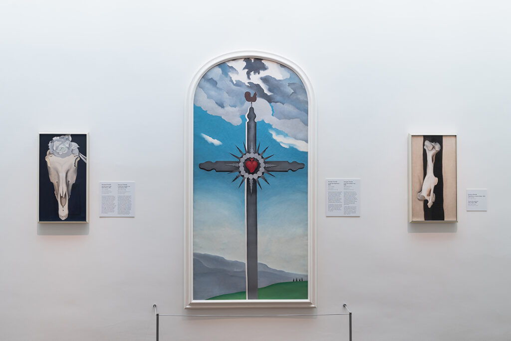 Photograph of a vertical painting of a grey cross with a red heart at its center. Behind the cross are green hills and blue sky. The top of the canvas is curved while its bottom is flat. The entire painting is in a white frame. To the left of the painting of the cross is a smaller vertical painting of a cow skull with a white flower on a black background. To the right of the painting of the cross is a smaller vertical painting of a thigh bone against a black strip and a beige background. All three paintings hang on the white wall of a Museum Gallery. In front of the cross painting is a small cordoned off area near the ground.