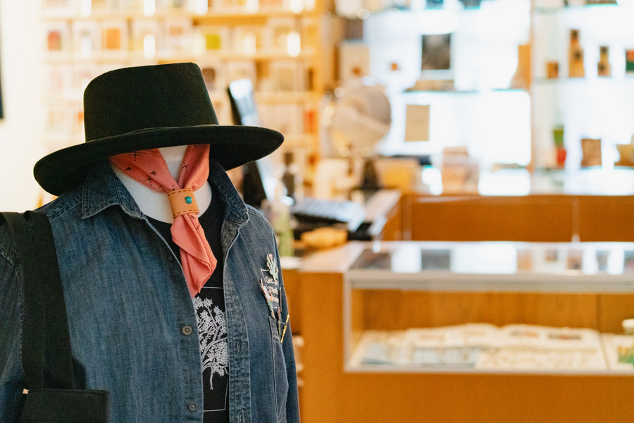 A mannequin with a black gaucho hat, a red tied scarf, a t-shirt, and a denim vest. Behind the mannequin in the background are shelves of the Museum Store.