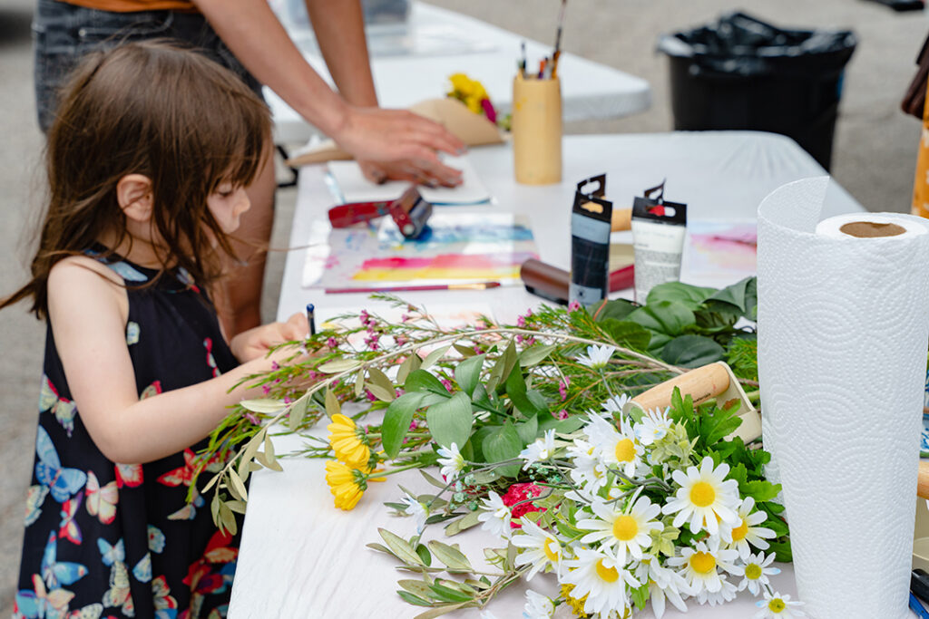 Photograph of a young child making an art piece on a table that is covered in art supplies and flowers.