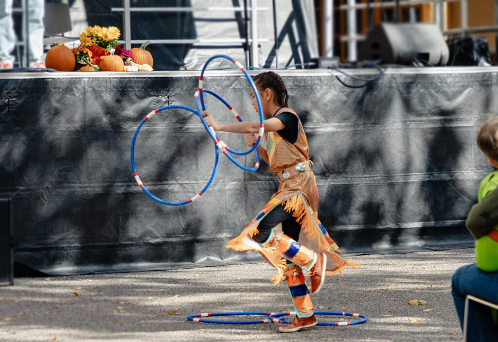 Photograph of a young child in traditional Tewa garb dancing with blue hoops around their ankles and in their hands. Behind the dancer is the dark edge of a stage.