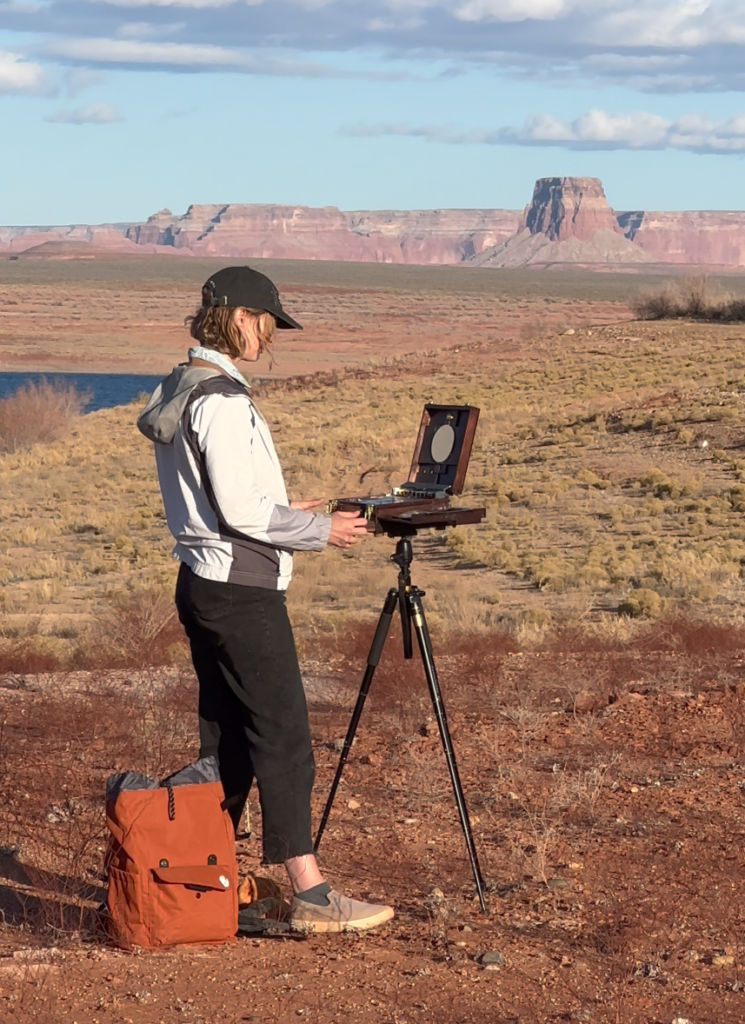 A person stands outdoors in an arid landscape with a mountain range in the background, painting on an easel. They wear a hat and jacket, and a backpack rests on the ground beside them.
