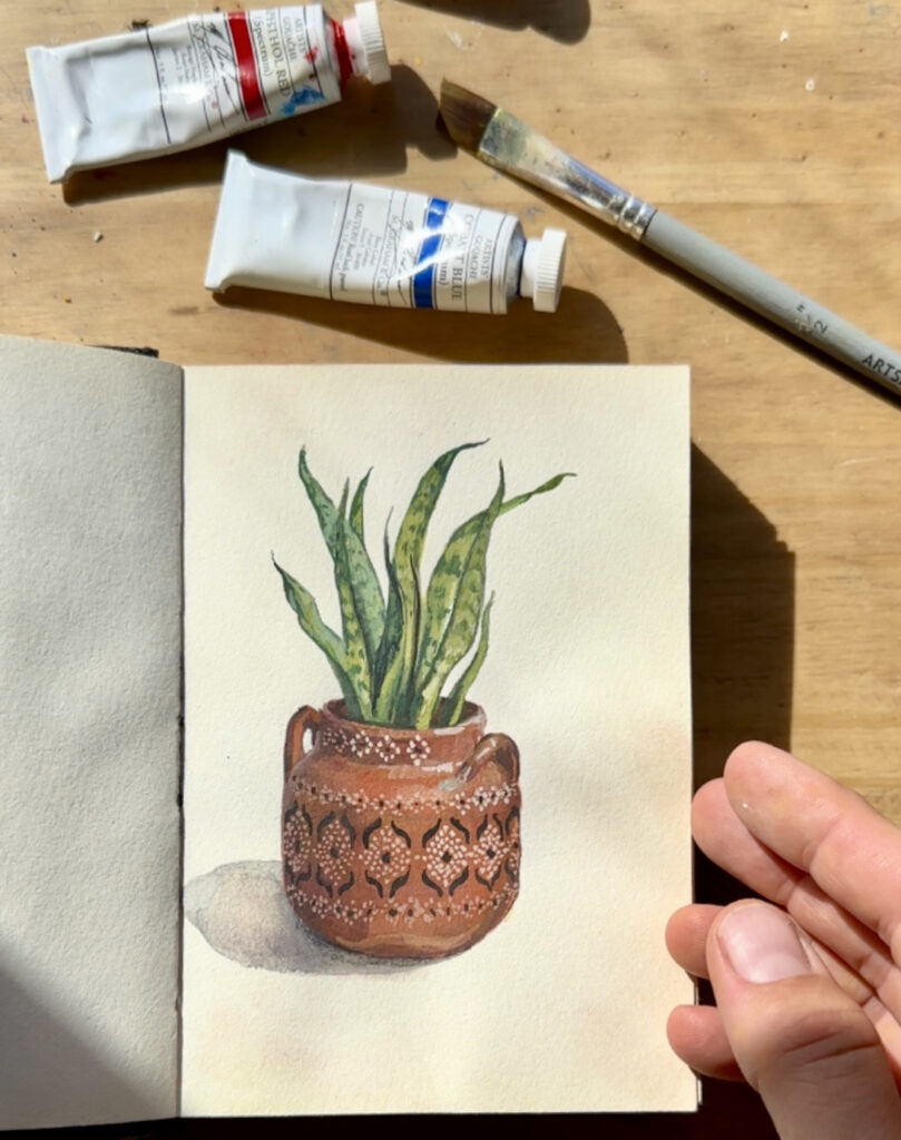 Photograph of a small book open to a page with a watercolor of a pot with a plant in it. To the right of the page a hand holds the book open. Above the book are small paint tubes and a paintbrush.