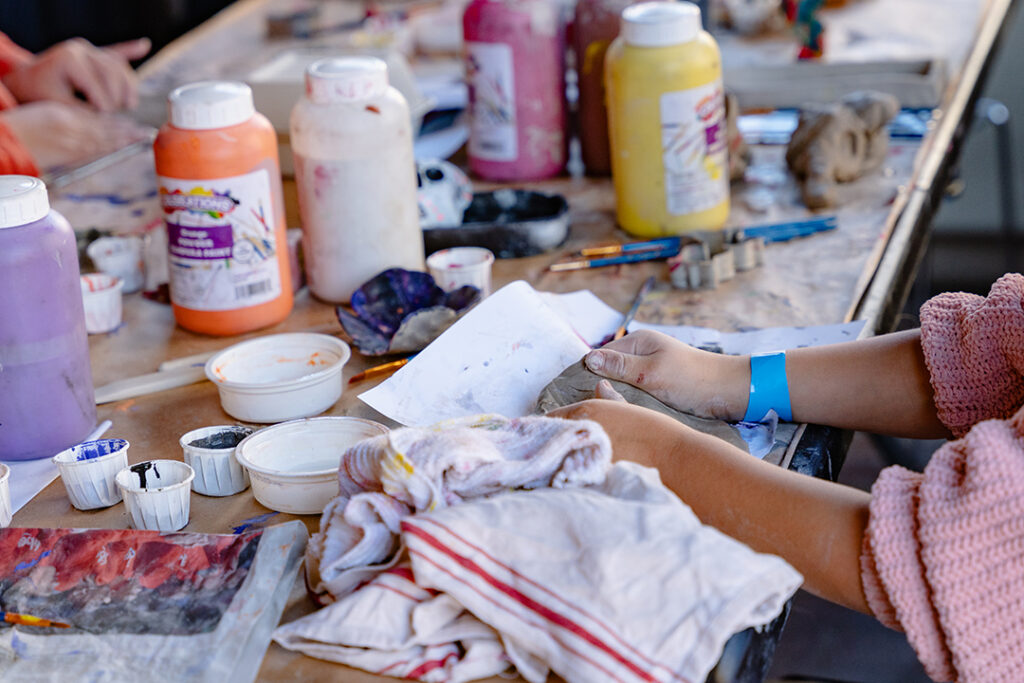 Close-up of a young person's hands as they mould clay on a table covered with paint and other art materials.