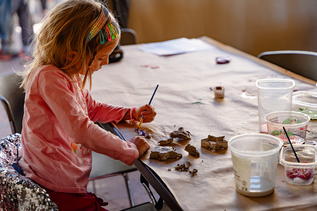 Photograph of a child sitting at a table making art with pieces of clay and a paintbrush.