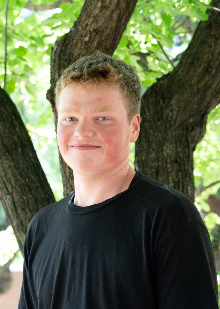 Photograph of a young person with short hair and a dark shirt smiling as they look in the camera. Behind them are green trees. 