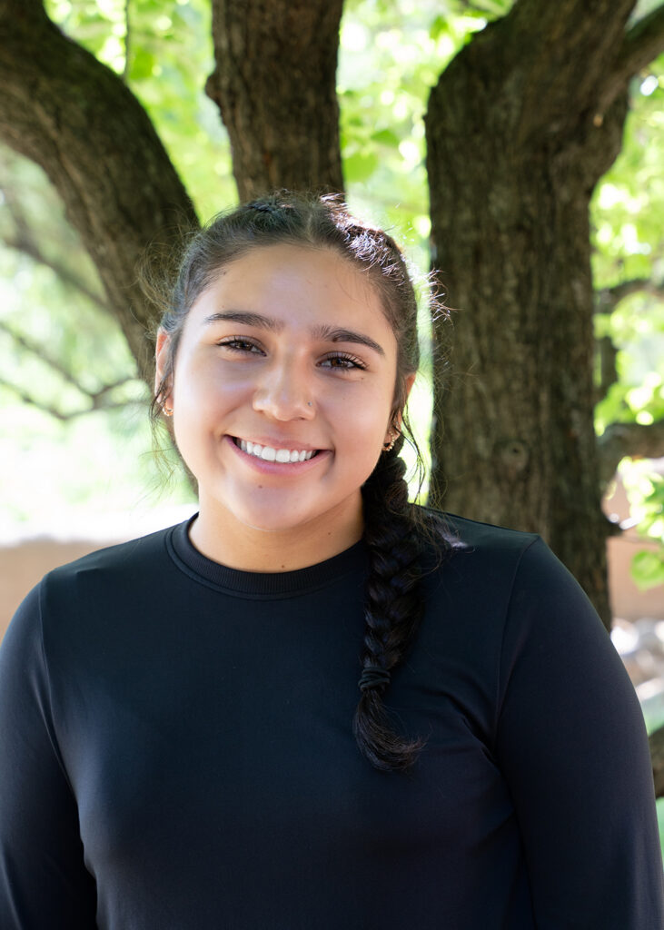 Photograph of a young person with long dark hair in two braids. They smile at the camera. Behind them are green trees. 