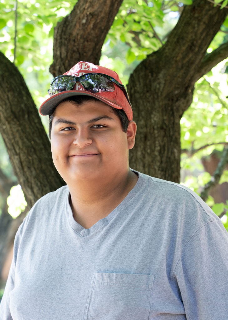 Photograph of a younger person with short hair wearing a hat and sunglasses perched on the hat. They are smiling at the camera and behind them are green trees. 