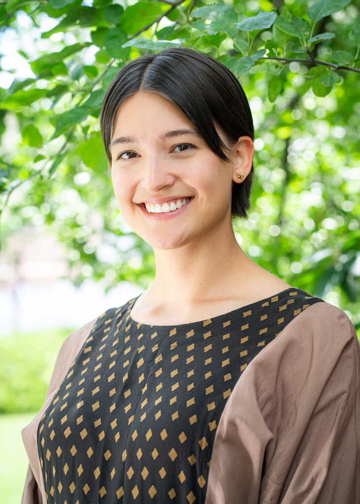 Photograph of a young person with short dark hair smiling at the camera. Behind them are green leaves and foliage.