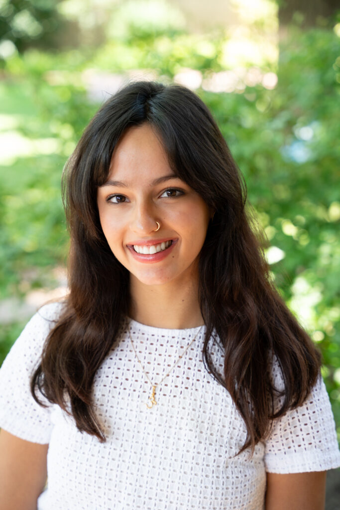 Photograph of a young person with long dark hair smiling at the camera. Behind them are green leaves and foliage.