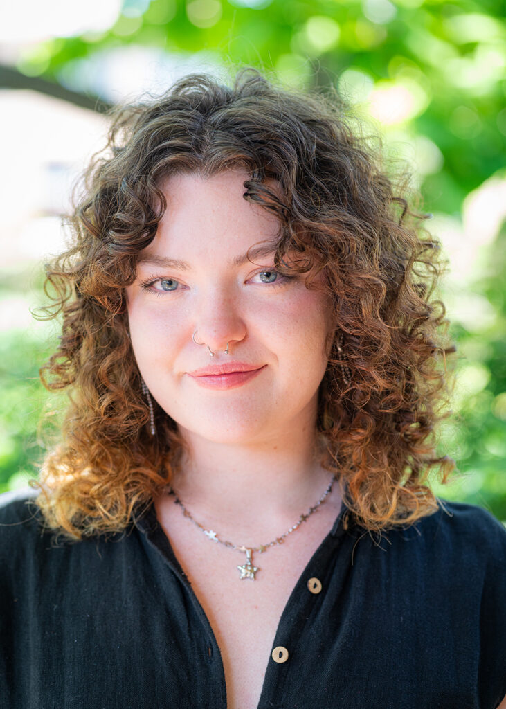 Photograph of a young person with shoulder-length curly hair earrings and necklaces. They stare into the camera with a slight smile. Behind them are green leaves and foliage.