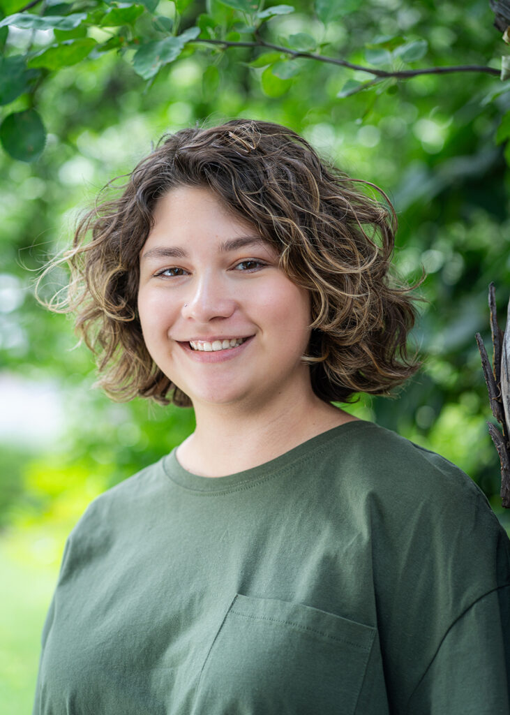 Photograph of a young person with curly short dark hair smiling at the camera. Behind them are green leaves and foliage.