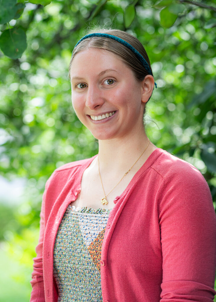 Photograph of a young person with hair tied up and a headband smiling at the camera. Behind them are green leaves and foliage.