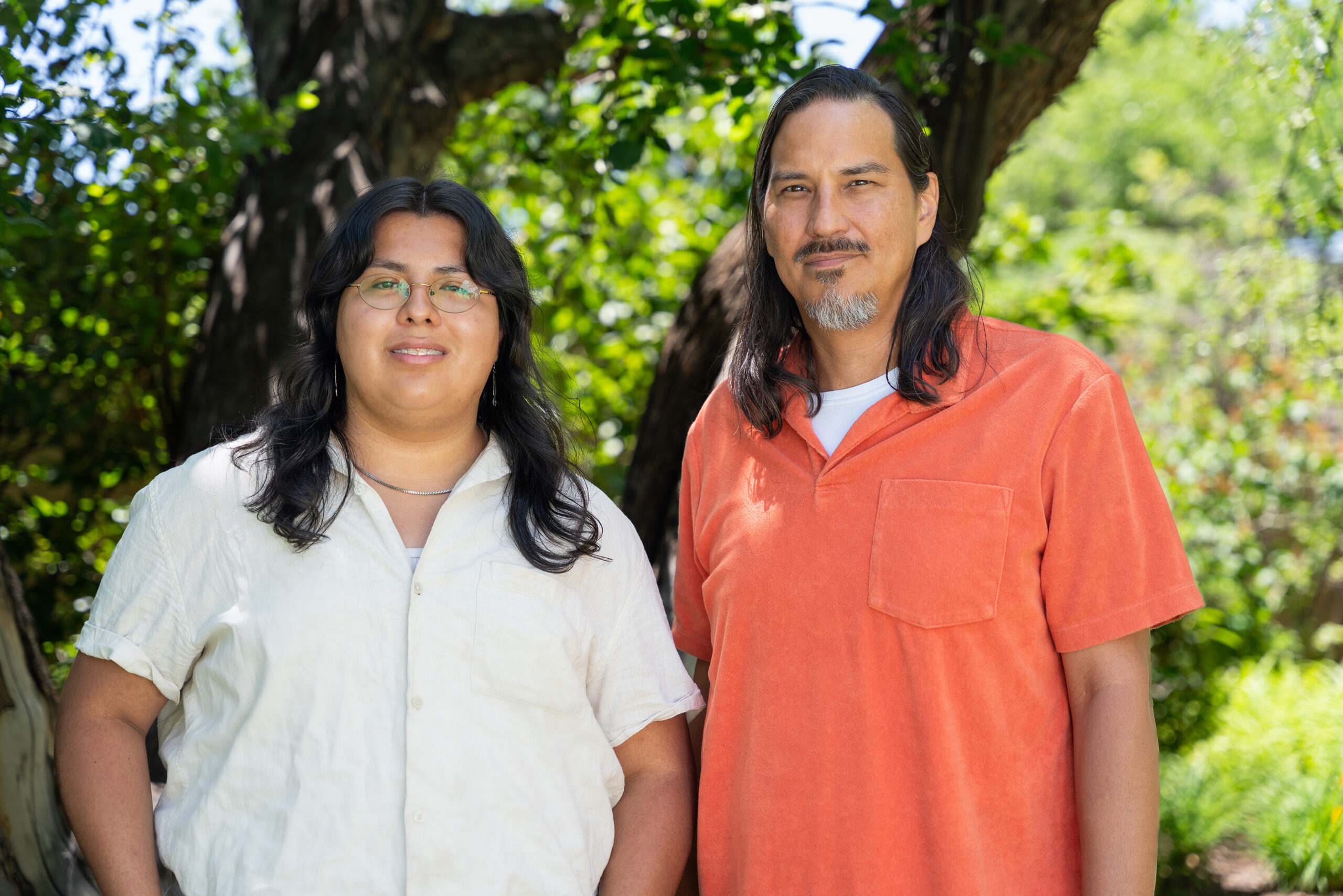 Photograph of two people standing side by side. The person on the left is shorter and wears glasses and a light button-up shirt. The person on the right is older and wear an orange shirt. Both look straight at the camera. Behind them are green trees