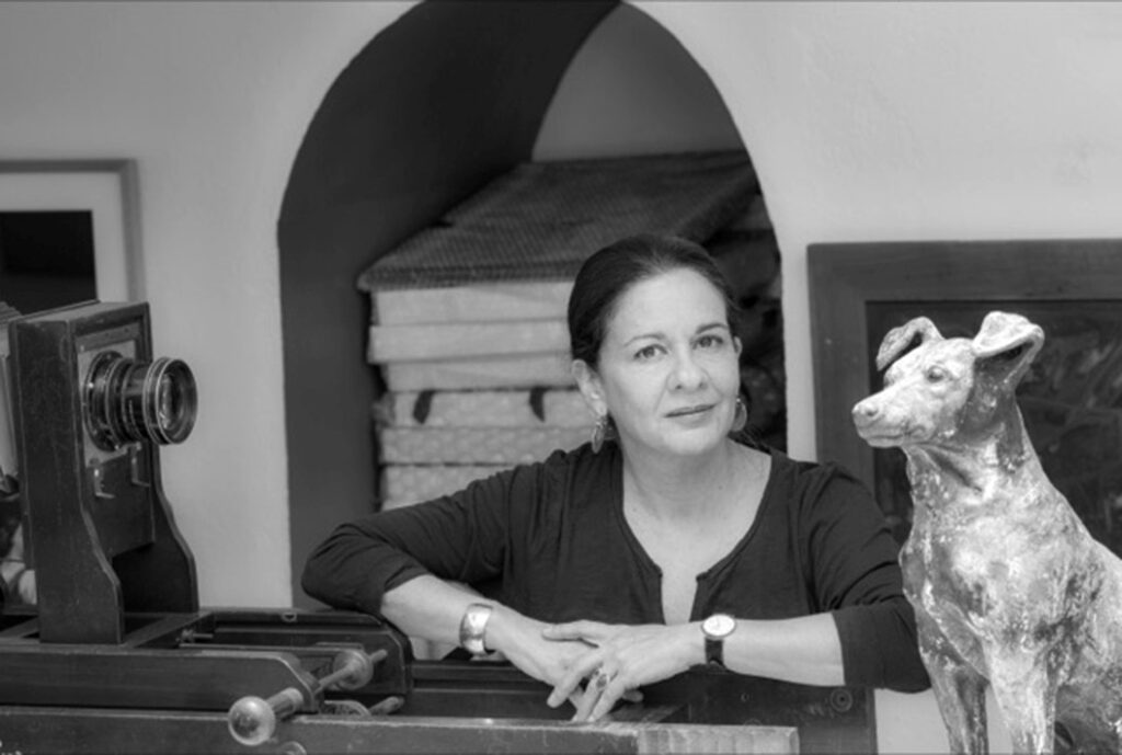 Black and white photograph of a person with their hands together resting on a table with a camera. Their hair is tied. To the right of the frame is a statue of a dog.