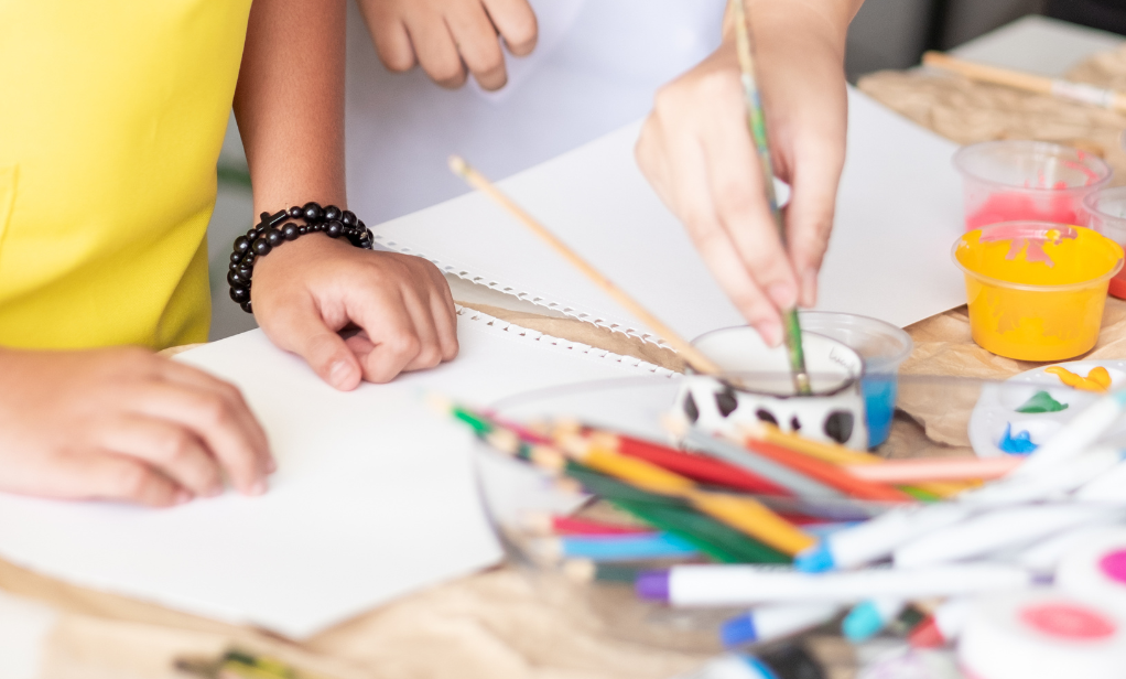 Close-up photograph of two young children's hands on a table as they paint and an adult's hands poke into view.