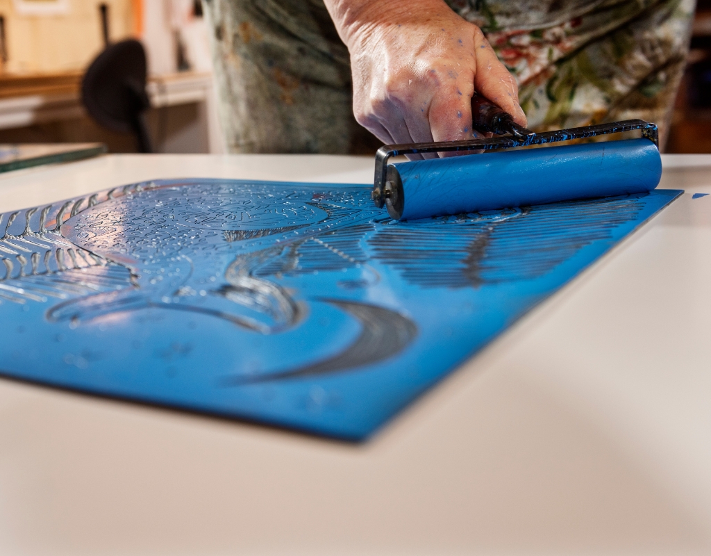 Photograph of a person's hand holding a brush as they spread blue ink on a carved material and prepare to make a print.