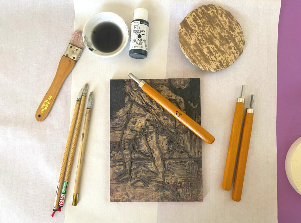 Photo of woodcut printmaking tools including brushes ink and knives on a white fabric. To the right is a purple sliver of fabric and at the center is block ready for print.