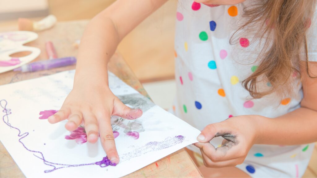 Photograph of a young child's hands as they use their fingers to paint a painting on a while piece of paper.