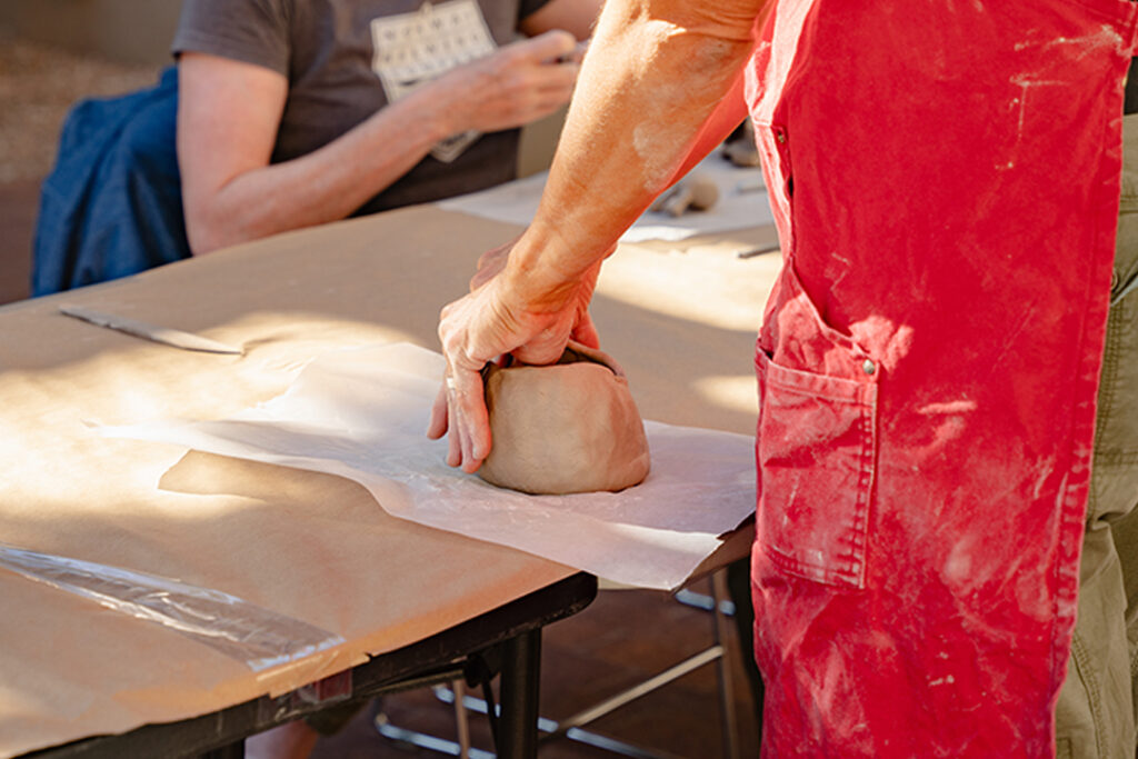 Photograph of a person from below the elbow wearing a red apron. The focus is on their hands as they shape a bowl made of clay at a able covered with a sheet of paper.