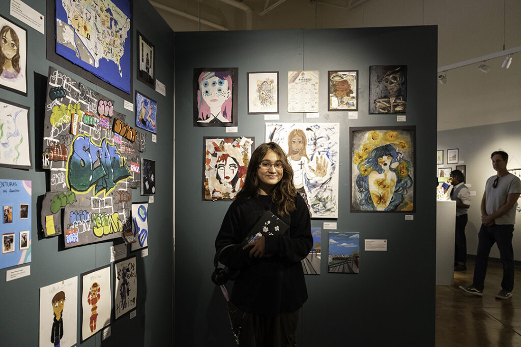 A young student with glasses and long hair stands in front of a gallery wall covered in art.