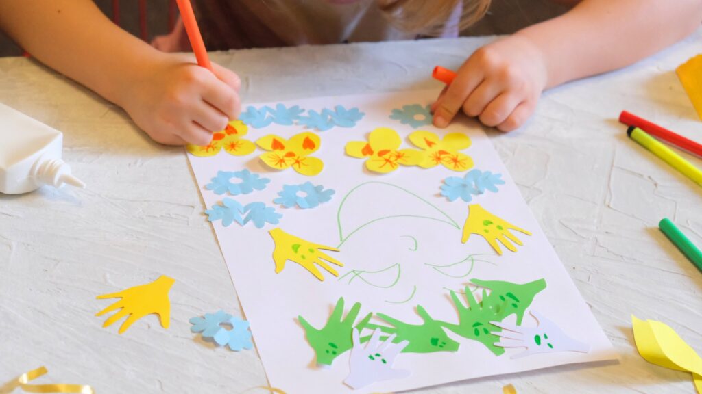 Close-up photograph of a young person's hands as they draw on a piece of paper covered with colorful cut-outs