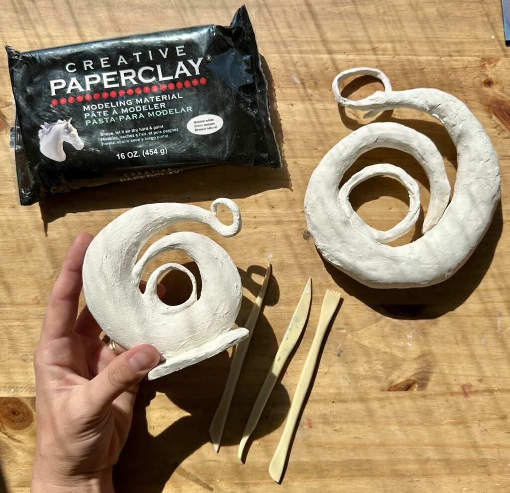 Photograph of two small rounded sculptures made of white paperclay laying on a wooden table. A hand holds the left swirling sculpture. Between the two are a set of small wooden tools. Above the hand is a packet of paperclay in black plastic packaging.