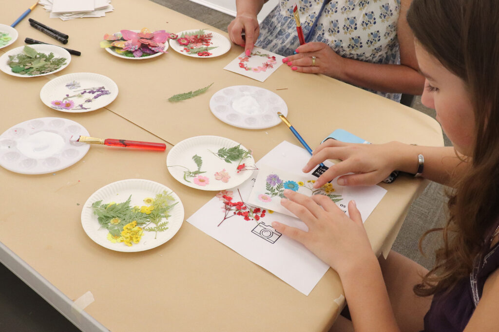 Photograph of a young person's hands as they glue flowers to a piece of paper on a table. To their right another person is pressing a brush against a piece of paper.