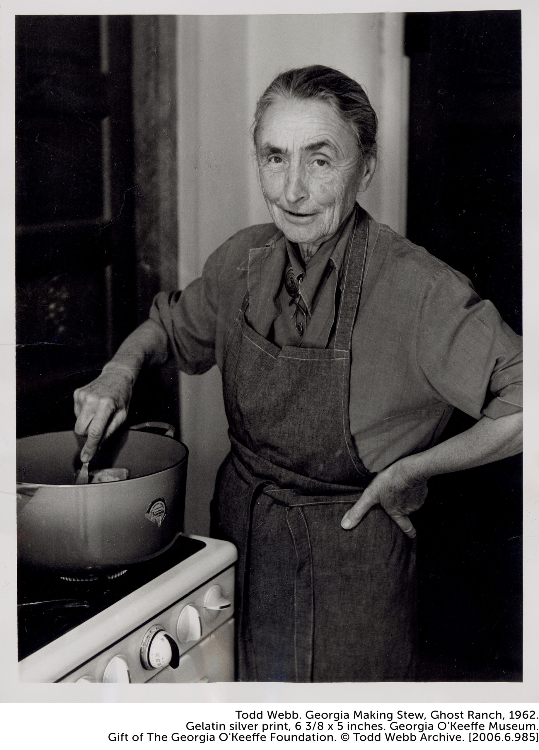 Portrait of O'Keeffe at the stove stirring stew, wearing a denim apron with one hand on her hip and the other hand stirring the pot.