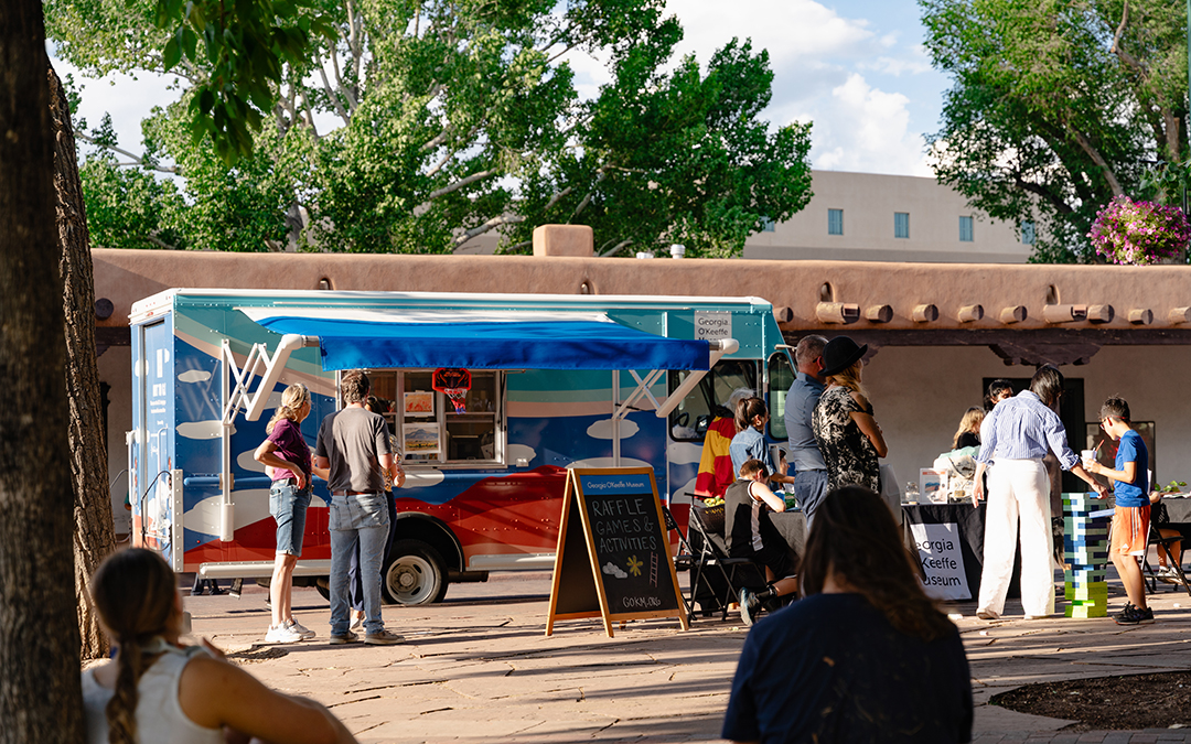 Photograph of the Art to G.O. Truck in shades of blue and red with people standing in front of it and a tree on the left side of the frame. 