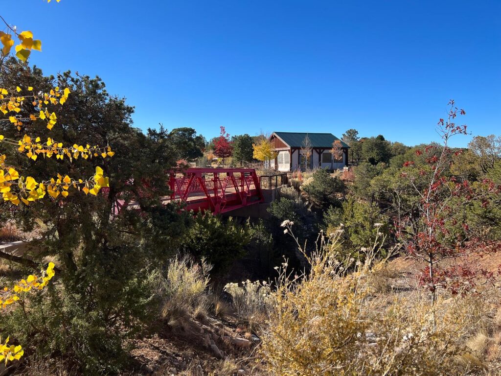 Photograph of a small red bridge leading to a wooden slant-roofed pavilion. The bridge and landscape is covered in green trees as well as some trees with orange and red leaves. Thy sky above is a deep blue.